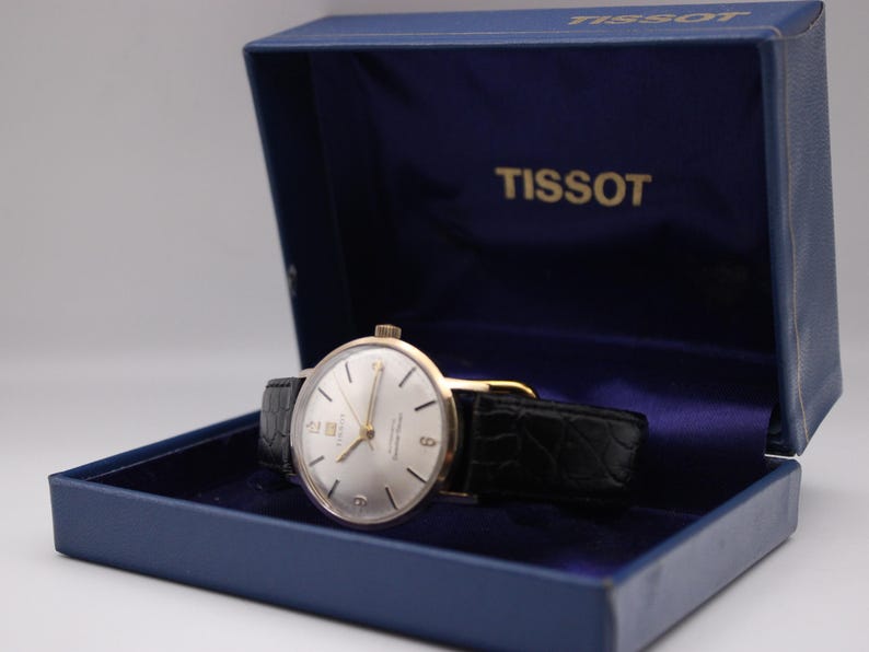 Może przedstawiać: Zegarek Tissot w stylu vintage ze srebrną tarczą i złotymi wskaz&oacute;wkami, umieszczony w niebieskim pudełku. Zegarek posiada czarny sk&oacute;rzany pasek. Otwarte pudełko eksponuje zegarek i nazwę marki "TISSOT".