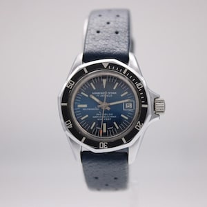 Può includere: Un orologio Marine Star vintage con quadrante blu e lunetta girevole nera. L'orologio ha una cassa argentata e un cinturino in pelle blu. Il quadrante mostra il testo "MARINE STAR 17 JEWELS SELF WINDING INC. BLOC WATER RESISTANT 66 FEET".