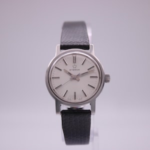 Puede incluir: Un reloj de pulsera Eterna de tono plateado con una esfera redonda blanca, marcadores de hora y manecillas plateadas. El reloj tiene una correa de cuero negro texturizado y una pequeña corona en el lado derecho de la caja. El logotipo Eterna está impreso.