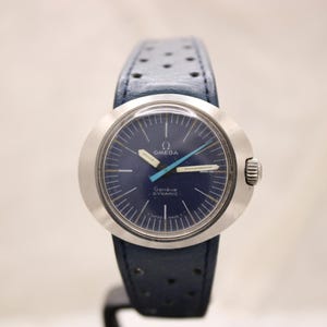 Pode incluir: Um relógio Omega Genève Dynamic vintage com mostrador azul marinho e caixa prateada. O relógio apresenta uma pulseira de couro azul com perfurações. O mostrador exibe o logotipo da Omega e as palavras "Genève Dynamic".
