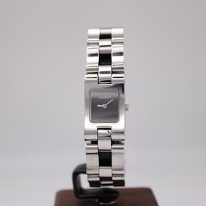 Puede incluir: Un reloj Gucci plateado con una esfera rectangular gris oscuro. El reloj tiene una pulsera de eslabones. La palabra "GUCCI" está impresa en la esfera. El reloj se muestra sobre un soporte negro.