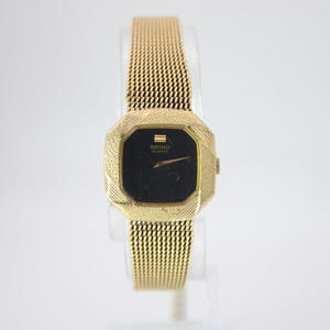 May include: A gold-toned Seiko Quartz watch with a black square face and a mesh strap. The watch has a textured bezel and a gold-toned mesh strap. The watch is displayed against a white background.