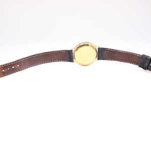 Back view of the Raymond Weil watch unbuckled, lying flat, revealing the underside of the black leather strap and the gold case back.