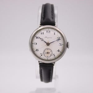 May include: A vintage Longines wristwatch with a white face, black hands, and a small seconds dial. The watch has a silver-toned case and a black leather band. The watch face displays the Longines logo.