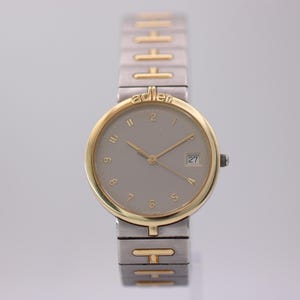 May include: A two-tone wristwatch featuring a round gold-tone bezel with the brand name adler at the top. The watch has a light gray dial with gold-tone numerals, thin hands, and a date window at the three o'clock position. The band is silver-tone metal with gold-tone H-shaped links.