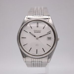 Könnte beinhalten: Eine silberfarbene Seiko Quartz-Uhr mit weißem Zifferblatt und Datumsanzeige auf 3 Uhr. Die Uhr hat ein silbernes Armband und ein rundes Zifferblatt mit schwarzen Zeigern und Markierungen. Auf dem Zifferblatt steht "SEIKO QUARTZ".