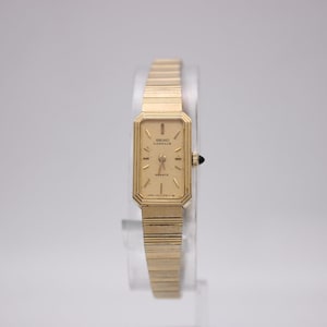 May include: A gold-toned Seiko Lassale quartz watch. The rectangular watch face has a gold dial with black hands and markers. The watch has a gold-toned, linked strap. The watch is displayed on a clear stand.