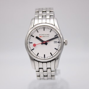May include: A silver-toned wristwatch with a white face, black hands, and a red second hand. The watch features a date display and a silver link band. The watch face displays the brand name "MONDAINE" and the text "SWISS SBB CFF FFS".