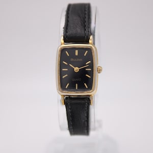 Puede incluir: Un reloj Bulova Quartz rectangular con esfera negra y caja dorada. El reloj tiene una correa de cuero negro. La esfera del reloj tiene agujas e indicadores dorados. El reloj se muestra sobre un fondo blanco.