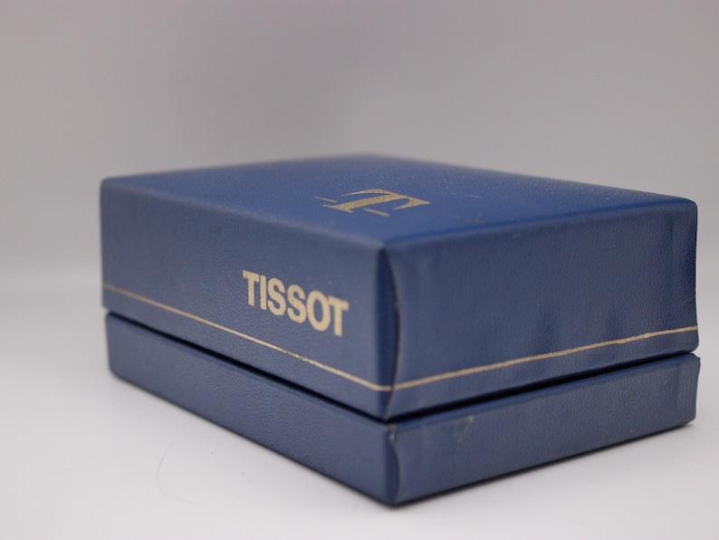 Może przedstawiać: Granatowe prostokątne pudełko ze złotym obramowaniem i nazwą marki "TISSOT" wydrukowaną na złoto. Pudełko ma złote logo na g&oacute;rze. Pudełko jest zamknięte i wydaje się być wykonane ze sk&oacute;ry.
