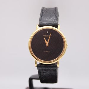 Front view of a gold Raymond Weil watch with black wave pattern dial and diamond at 12 o'clock, on a black leather strap.