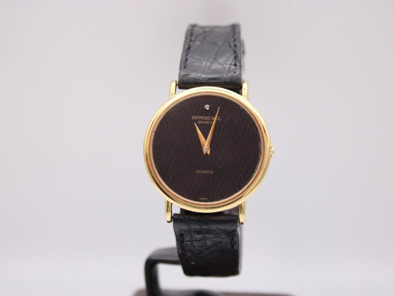 Front view of a gold Raymond Weil watch with black wave pattern dial and diamond at 12 o'clock, on a black leather strap.