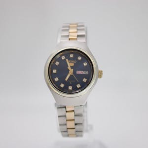 May include: A silver and gold-toned Seiko watch with a dark blue face. The watch features diamond-like markers and gold hands. The watch band is a combination of silver and gold links. The day and date are displayed at the 3 o'clock position.