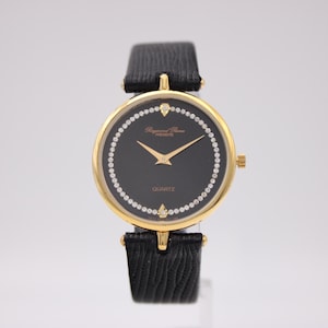 Puede incluir: Un reloj de pulsera dorado con esfera negra y bisel con diamantes. El reloj tiene agujas doradas y una correa de cuero negro. La esfera indica "QUARTZ".