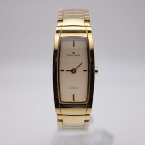 May include: A gold-toned Marco Coreani wristwatch with a rectangular face and a matching gold band. The watch face is a light champagne color with black hands and markers. The watch is water resistant.