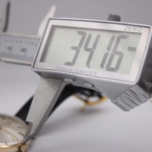 Może przedstawiać: Cyfrowa suwmiarka mierzy złoty zegarek na rękę. Wyświetlacz pokazuje 34,16 mm. Zegarek ma złotą kopertę, białą tarczę i czarny sk&oacute;rzany pasek. Suwmiarka jest srebrna.