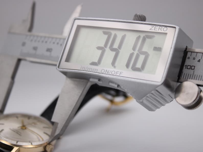 Może przedstawiać: Cyfrowa suwmiarka mierzy złoty zegarek na rękę. Wyświetlacz pokazuje 34,16 mm. Zegarek ma złotą kopertę, białą tarczę i czarny sk&oacute;rzany pasek. Suwmiarka jest srebrna.