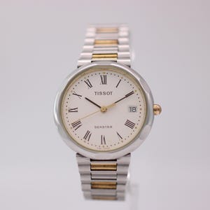 May include: A Tissot Seastar wristwatch featuring a round silver-tone case and a white dial with black Roman numeral hour markers. The watch includes a date window at the three o'clock position and a two-tone silver and gold-tone metal link bracelet.