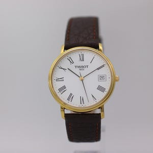 May include: A Tissot 1853 wristwatch with a round gold-tone case, a white dial featuring black Roman numeral hour markers, thin black hands, and a date window at the three o'clock position. The watch has a textured dark brown leather strap.