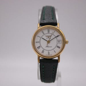 Puede incluir: Un reloj Longines Presence de cuarzo, dorado, con esfera blanca y números romanos negros. El reloj tiene una ventana de fecha a las 3 en punto y una correa de cuero negro con costuras verdes. La esfera del reloj dice "Longines Quartz Presence".