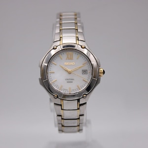 May include: A Seiko Coutura wristwatch with a silver and gold-tone stainless steel band and case. The watch has a white face with gold-tone hands and markers, and a date display at the 3 o'clock position.