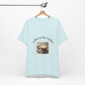 May include: Pale blue t-shirt with the text "Coffee is My Copilot" in a curved font. The shirt has a small image of a coffee cup with latte art. The t-shirt is hanging on a wooden hanger.