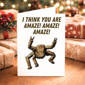 Könnte beinhalten: Eine weiße Grußkarte mit dem Text "I THINK YOU ARE AMAZE! AMAZE! AMAZE!" in Braun. Die Karte zeigt eine Cartoon-Illustration einer braunen, felsartigen Kreatur, die Friedenszeichen macht. Der Hintergrund umfasst verpackte Geschenke.