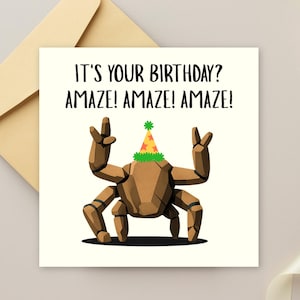 May include: A square greeting card featuring a brown, multi-legged rock creature wearing a colorful party hat with a green fringe. The text reads: IT'S YOUR BIRTHDAY? AMAZE! AMAZE! AMAZE! The card is positioned against a light-colored envelope.