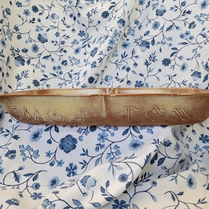 May include: A rectangular, divided serving dish with a light brown and tan glaze. The dish has embossed symbols and numbers on the front. The background is a white fabric with a blue floral pattern.