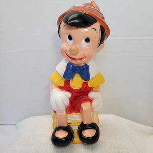 Może przedstawiać: Figurka Pinokia w stylu vintage. Postać ma na sobie brązowy kapelusz, biały kołnierz, niebieską muszkę, żółtą koszulę i czerwone ogrodniczki. Figurka siedzi na stosie książek i ma czarne buty.