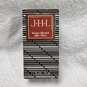 May include: A rectangular box of J.H.L. Custom Blended After Shave. The box has a black and white striped pattern. The front of the box features a red and gold label with the text "J.H.L." and "Custom Blended After Shave". The box contains 1.7 fl. oz. / 50 ml.