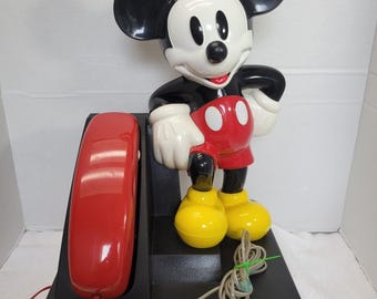 Pode incluir: Um telefone vintage do Mickey Mouse. O telefone apresenta um auscultador vermelho, uma base preta e uma grande figura do Mickey Mouse com sua roupa clássica. A figura é preta, branca, vermelha e amarela.