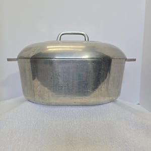 May include: A large, oval-shaped, aluminium cooking pot with a matching lid. The pot has a handle on the lid and two side handles. The metal has a brushed finish and shows signs of wear. The pot is designed for cooking and food preparation.