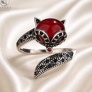 Op de afbeelding: Een zilverkleurige open ring in de vorm van een vos, voorzien van een grote, gladde, dieprode cabochonsteen als kop, geaccentueerd met kleine zwarte steentjes langs de oren en staart, en een enkel minuscuul rood steentje op de neus, met gravures.