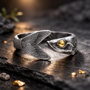 May include: A silver-toned ring shaped like a fish, featuring a detailed tail and head. The fish's eye is a contrasting gold color. The ring is designed to wrap around the finger, creating a unique and aquatic-themed accessory.