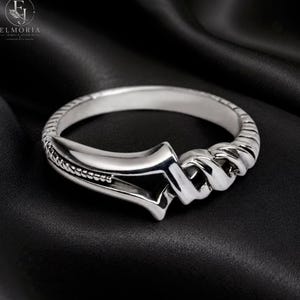 May include: A polished silver ring featuring a smooth, tapered band that transitions into a twisted, rope-like texture on one side. The design includes a central open geometric element and a beaded detail along the edge of the shank.