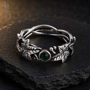 May include: A silver-toned ring featuring a band designed with intertwined metal vines and sculpted leaves. The ring is accented with a single, round, dark green gemstone set within the leaf pattern, creating a detailed botanical appearance.