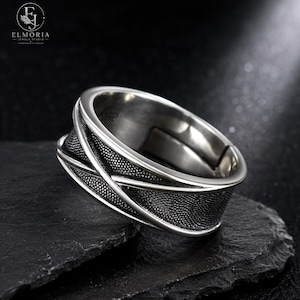 May include: A silver-toned metal ring featuring a wide band with a textured, dark-finished center section and smooth, polished edges. The design includes raised, crossing diagonal lines that create a geometric pattern across the surface of the band.