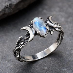 May include: A silver ring featuring an oval-shaped iridescent moonstone set between two crescent moon motifs. The band is crafted with a textured, branch-like design that wraps around the finger, showcasing oxidized silver details and highlights.