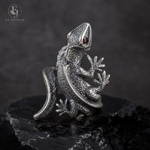 May include: A silver ring shaped like a lizard with a textured, scaly surface and a small, round, dark red gemstone set as the eye. The lizard's body curves to form the band of the ring, with its tail extending outward and its feet positioned.