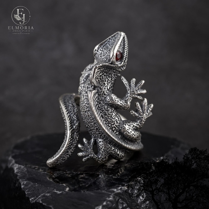 May include: A silver ring shaped like a lizard with a textured, scaly surface and a small, round, dark red gemstone set as the eye. The lizard's body curves to form the band of the ring, with its tail extending outward and its feet positioned.
