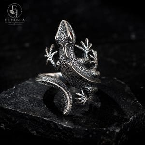 May include: A silver ring featuring a detailed, textured lizard design that wraps around the finger. The lizard has a dark, oxidized finish with visible scales and small red eyes. The ring is branded with the text ELMORIA JEWELS STUDIO.
