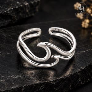 May include: A silver-colored open ring featuring a stylized wave design. The band has a polished metallic finish and includes the engraving S925 on the inner surface. The ring is adjustable with a gap between the two ends of the wave motif.