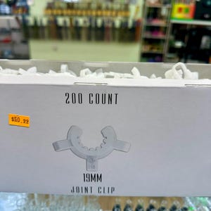 May include: A white box displaying "200 COUNT" and a 19mm joint clip graphic. The box has a yellow price tag. The background shows a shop display with various items.