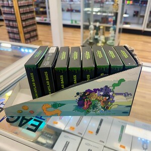 May include: A display of black boxes with green accents, labeled "Mylar Bags," arranged in a decorative cardboard container. The container features a colorful cartoon design. The boxes are on a glass display case.
