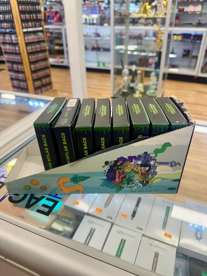May include: A display of black boxes with green accents, labeled "Mylar Bags," arranged in a decorative cardboard container. The container features a colorful cartoon design. The boxes are on a glass display case.