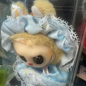 May include: A plush doll with a light brown face, large black eyes, and red lips. The doll wears a light blue dress with white lace trim and a matching bonnet. The text "FALL IN WILD" is visible on the packaging.