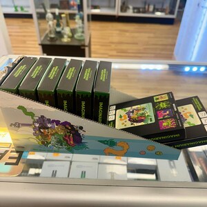May include: Display of MAGINARIUM DESIGNER SERIES boxes in a retail setting. The boxes are black with green accents and text. A colorful display with illustrations is in the foreground. The background shows glass display cases and store shelving.