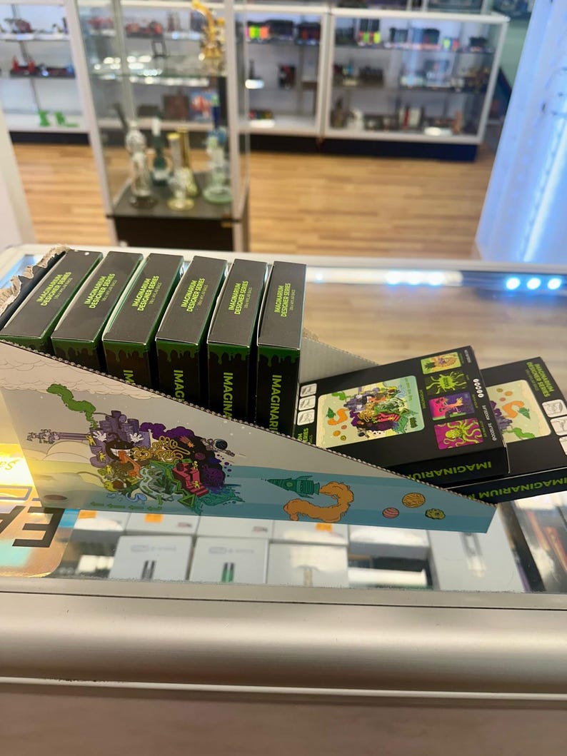 May include: Display of MAGINARIUM DESIGNER SERIES boxes in a retail setting. The boxes are black with green accents and text. A colorful display with illustrations is in the foreground. The background shows glass display cases and store shelving.