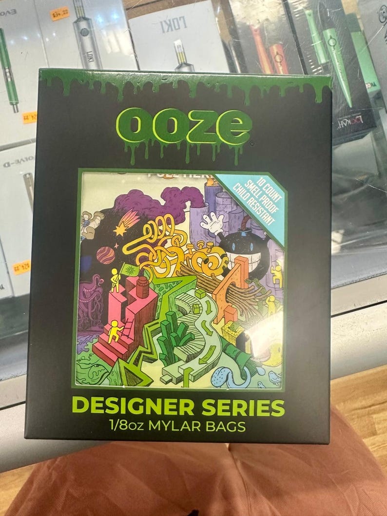 May include: Black box with the word "ooze" in green dripping letters. The box features a colorful cartoon illustration and text that reads "Designer Series 1/8oz Mylar Bags." The box also states "10 Count Smell Proof Child Resistant."